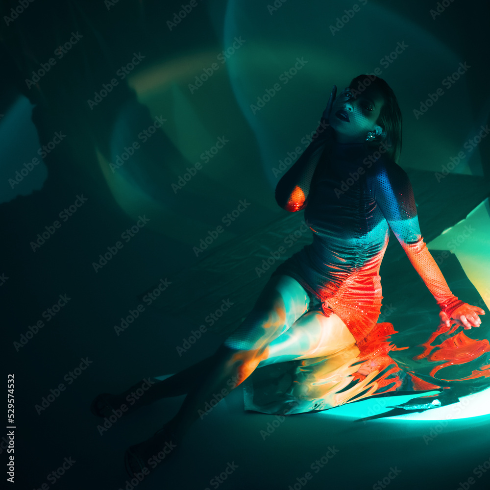 Glamorous portrait of a young beautiful blonde in a stylish scarlet short dress, sitting on the floor, refraction effects of colored blue light, futuristic fashion shooting in neon light