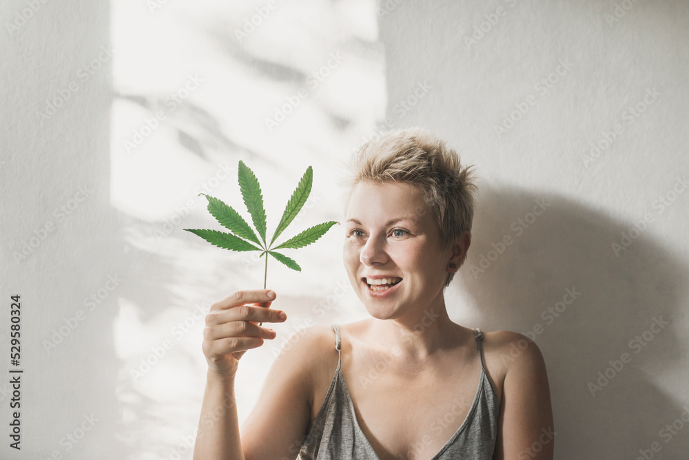 Blonde bohemian young woman holds a marijuana leaf in a close up cropped face portrait on shadows grey background. Short cut blonde Cannabis medicine, healthy lifestyle and ecology concept Blonde bohemian young woman holds a marijuana leaf in a close up cropped face portrait on shadows grey background. Short cut blonde Cannabis medicine, healthy lifestyle and ecology concept