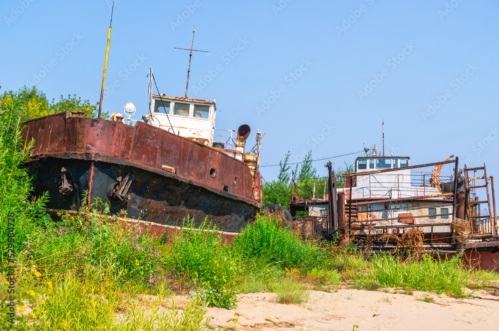 Rusty river ships lie on the sandy shore. Abandoned ships against a background of green bushes and blue sky. A rusting tugboat on the riverbank. Vessels that have served their service life.