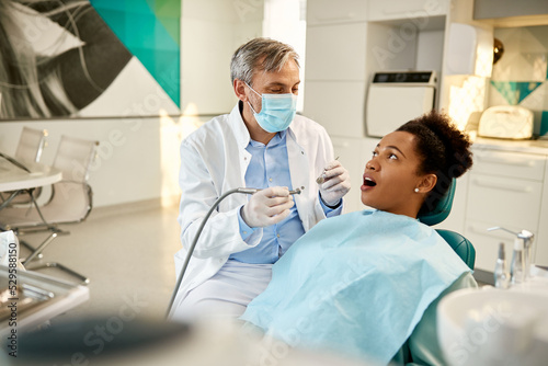 Dentist talks to black female patient during dental procedure at dentist s office.