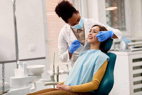 Black dentist wearing face mask while examining teeth of young woman at dental clinic.