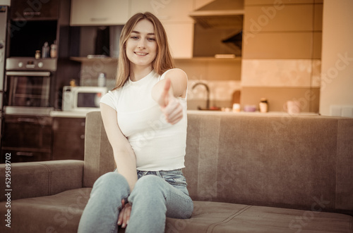 Young smiling woman showing thumb up while sitting on sofa in living room