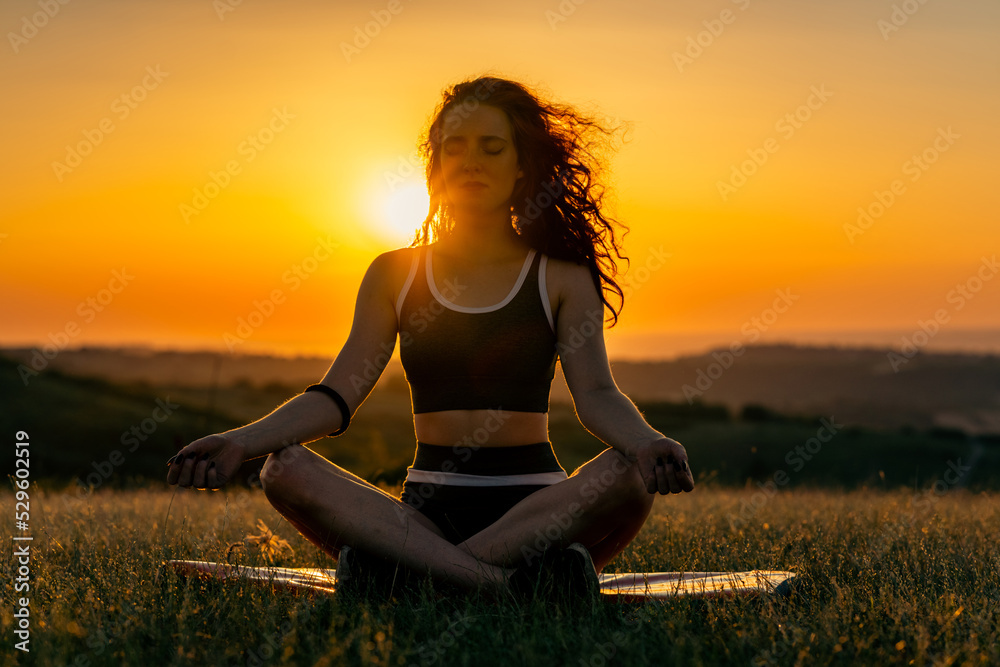 A young woman in shape practices meditation on yoga mats in the background of the setting sun and the layers of the mountains. Exercise and train outdoors. A sporty woman who practices yoga in nature. A young woman in shape practices meditation on yoga mats in the background of the setting sun and the layers of the mountains. Exercise and train outdoors. A sporty woman who practices yoga in nature.