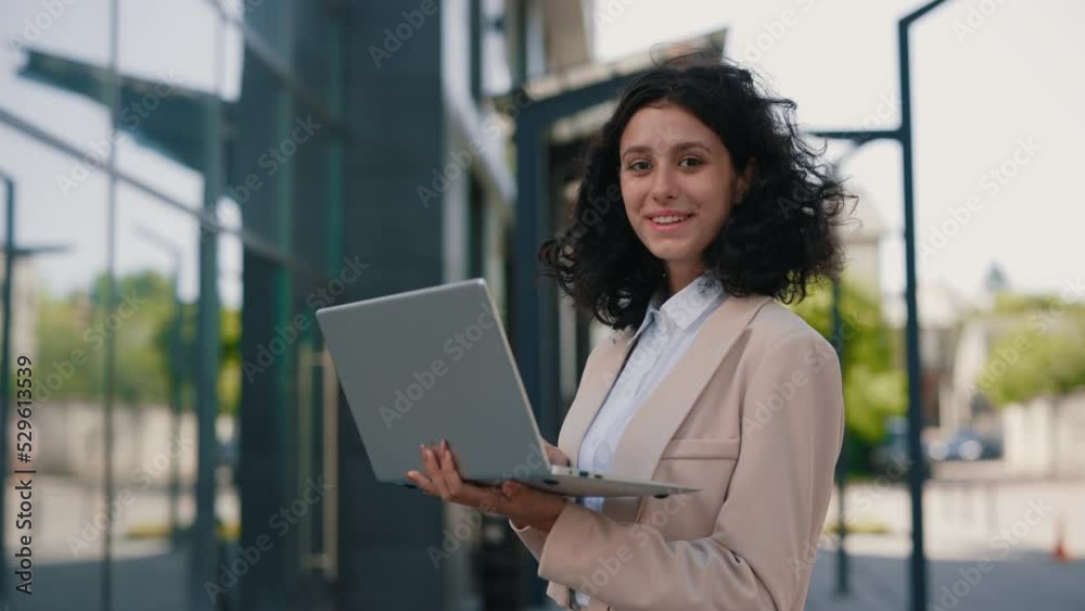 Portrait of the beautiful curly smiling young businesswoman in formal outfit standing near the business centre looking at the camera holding and using laptop for work Portrait of the beautiful curly smiling young businesswoman in formal outfit standing near the business centre looking at the camera holding and using laptop for work
