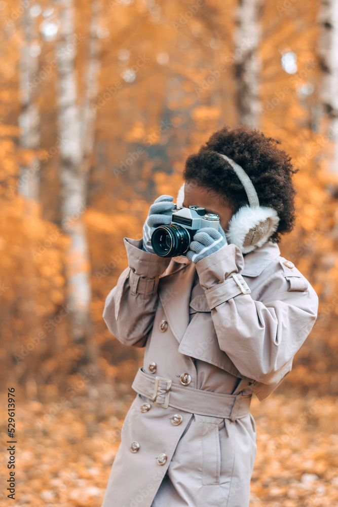 Cute African-American girl in fur headphones takes pictures with a camera in an autumn park.Diversity,autumn concept. Cute African-American girl in fur headphones takes pictures with a camera in an autumn park.Diversity,autumn concept.