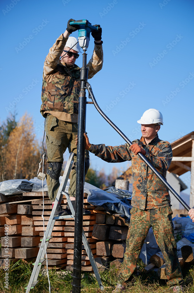 Male workers building pile foundation for wooden frame house. Men builders in white safety helmets drilling piles into the ground on blue sky background.