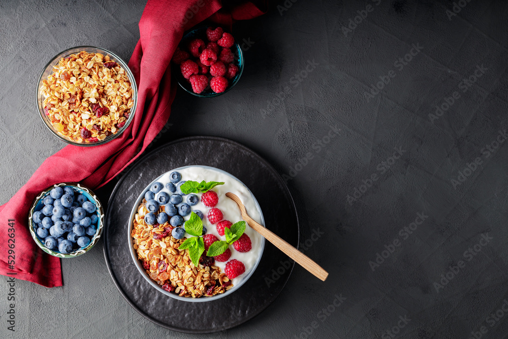 Breakfast cereal granola with berries and greek yogurt on a dark background. Bowl with yogurt, granola and fresh berries on a black plate. Healthy breakfast with wooden spoon