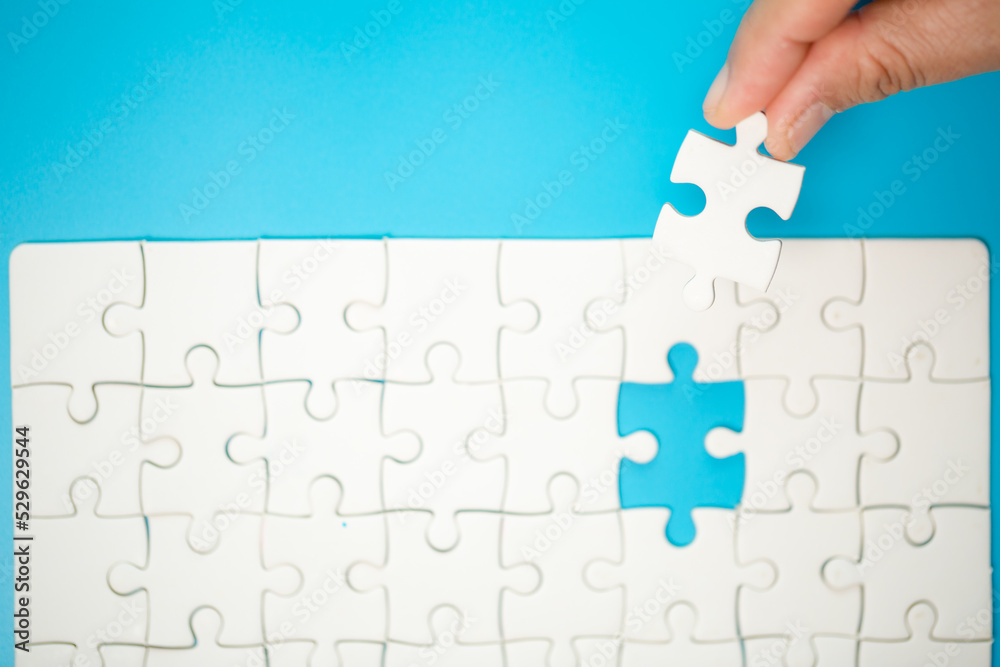 Close up hand holding and playing jigsaw game incomplete. White part of jigsaw puzzle pieces on blue background. concepts of problem solving, business, teamwork. Close up hand holding and playing jigsaw game incomplete. White part of jigsaw puzzle pieces on blue background. concepts of problem solving, business, teamwork.
