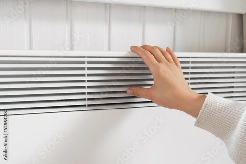 Hands in winter gloves warming up over electric heater. Frozen woman wearing a knitted mittens freezing for winter cold. Discomfort spending time at home. Girl warming hands on modern radiator.