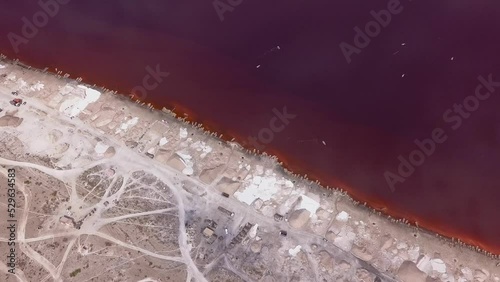 Aerial footage of stacks of salt and roads made of salt along a pink lake, clouds reflecting on the lake, Retba lake, Dakar, Senegal, facing down at the shore and going towards it while tilting down. photo