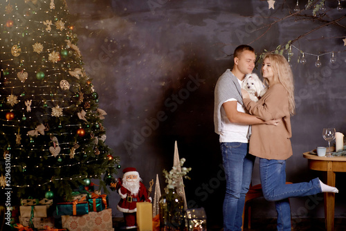 Portrait of beautiful family near Christmas tree. Romantic couple with their dog near in festive aesthetic cozy home interior. Good mood, Christmas love story. Candid, true moment.