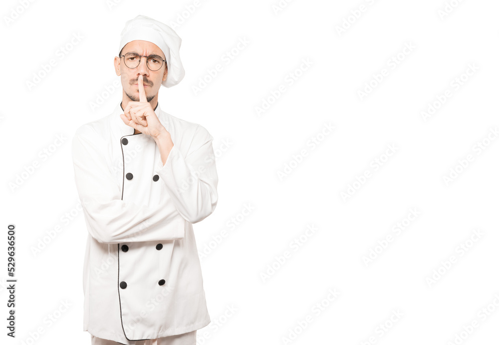 Serious young chef asking for silence gesturing with his finger Serious young chef asking for silence gesturing with his finger