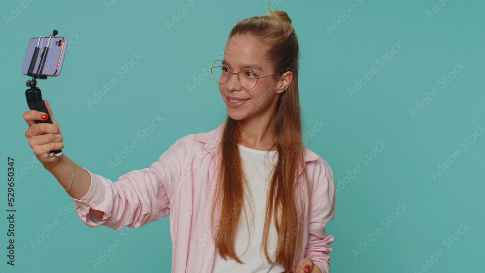 Pretty teenager girl traveler blogger taking selfie on mobile phone selfie stick, communicating video call online with subscribers. Young child kid isolated alone on blue studio background indoors Pretty teenager girl traveler blogger taking selfie on mobile phone selfie stick, communicating video call online with subscribers. Young child kid isolated alone on blue studio background indoors