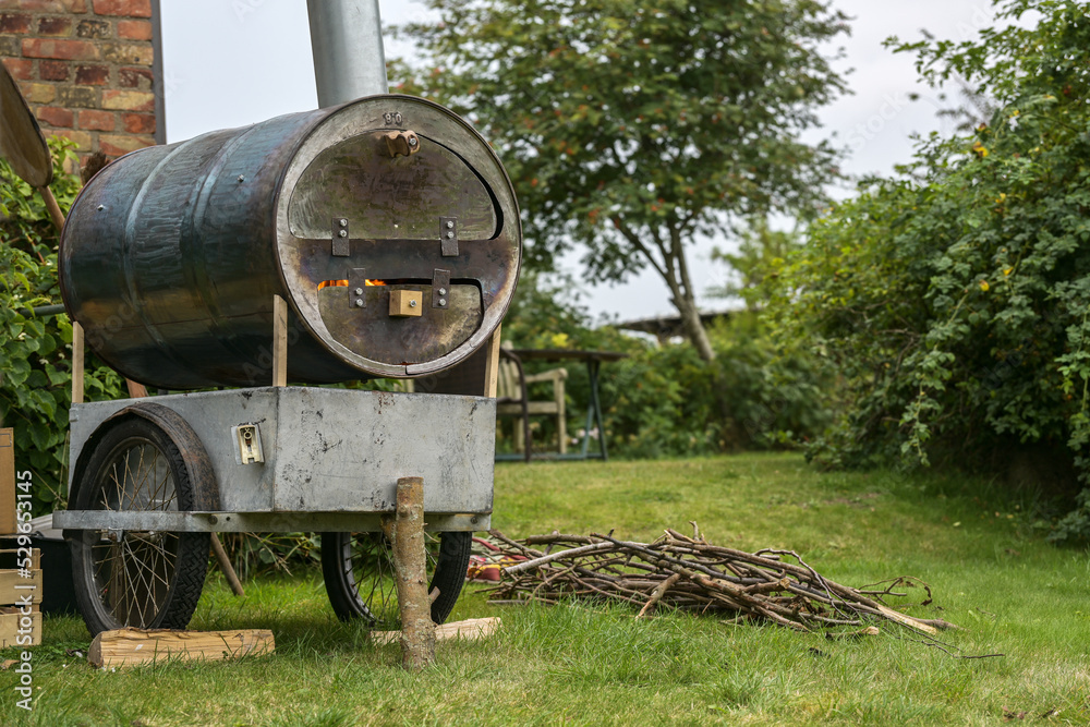 Homemade mobile pizza oven from an old metal barrel on a recycled chassis in a garden, fire in the combustion chamber and firewood on the lawn, copy space Homemade mobile pizza oven from an old metal barrel on a recycled chassis in a garden, fire in the combustion chamber and firewood on the lawn, copy space