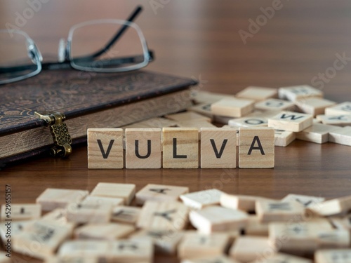 vulva word or concept represented by wooden letter tiles on a wooden table with glasses and a book