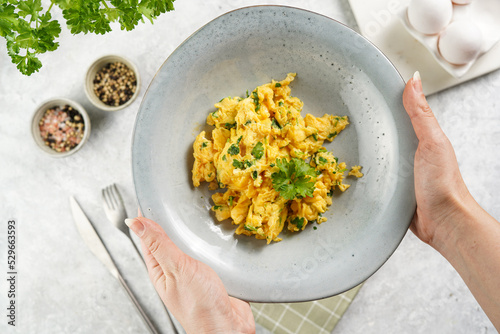 Female hands holding breakfast scrambled eggs with green herbs, parsley in deep grey bowl on grey neutral table, vintage fork and knife, top view
