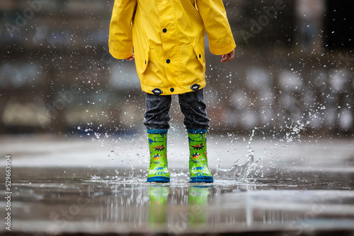 The child's yellow raincoat and green boots fall into a puddle of water. Autumn rainy weather. A walk in the rain.