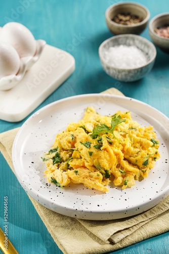 Breakfast scrambled eggs with green herbs, parsley in white plate on vivid blue wooden table, golden fork and knife