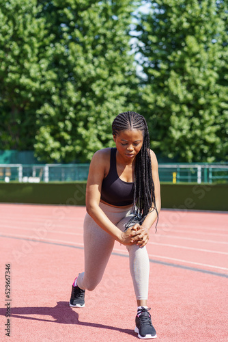 African American lady with long braids tries hard to pump gluteal muscles on red track of city stadium against lush green trees. Persistent woman takes care of fit body near park on summer day © DimaBerlin
