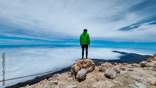 Rear view on hiking man in green wind jacket standing on rock on summit of volcano Pico del Teide on the island of Tenerife, Canary Islands, Spain, Europe. Island covered in cloud. Freedom concept