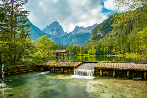 Famous green lake Schiederweiher near village Hinterstoder. Small weir and dam on lake with flowing water. Big Austrian mountains in background. Soft and magical colors in pure nature. © Martin