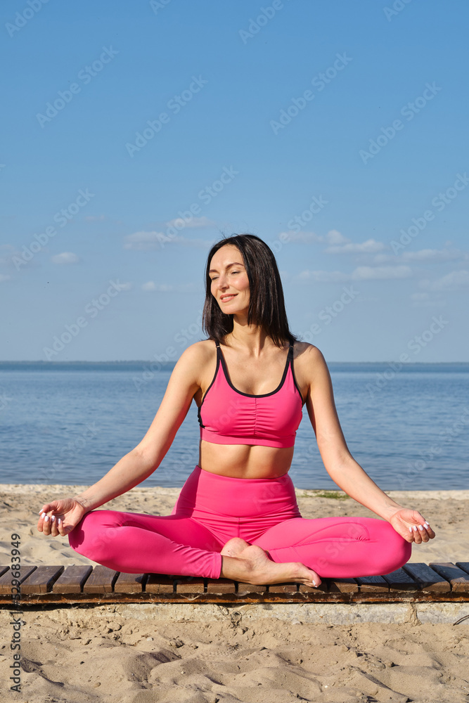Women in sportswear sitting in a lotus position and meditating during yoga classes on the seashore. Practicing yoga lesson, breathing, meditation, doing Ardha Padmasana exercises. Wellness concept