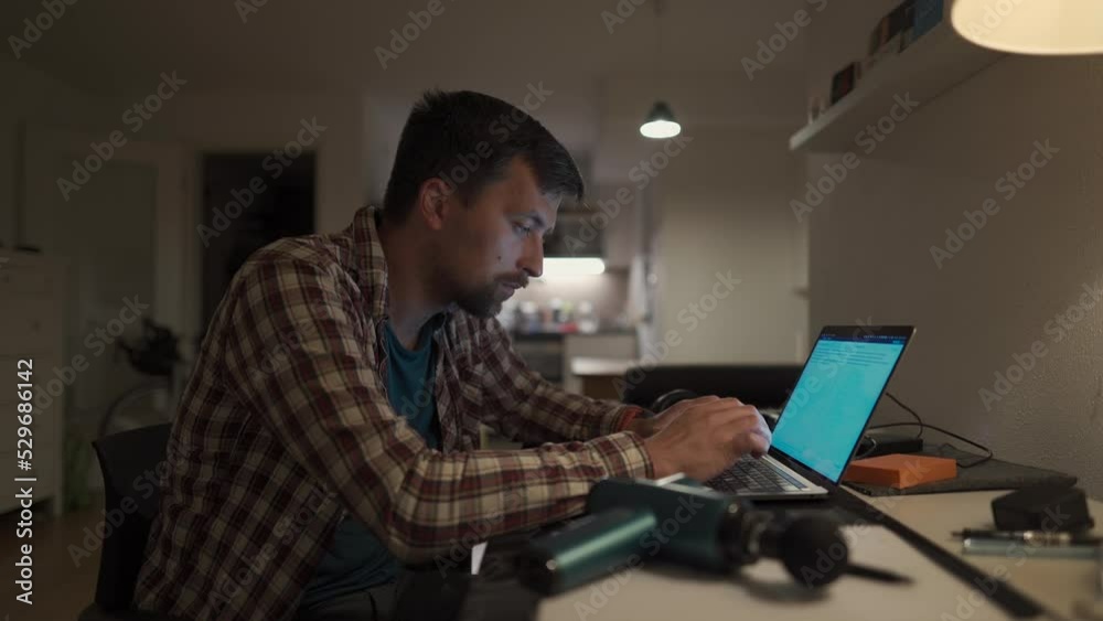 Tired male student studying on laptop at home until late and has neck pain and massages himself with percussion gun. Neck and back pain. Overworked man uses pistol massager to relieve muscle tension 