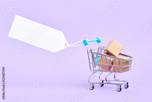 Shopping cart full of boxes with a blank label on a pastel lilac background. Minimalist design with copy space. Concepts: market deals, seasonal sales and discounts, black friday advertising.