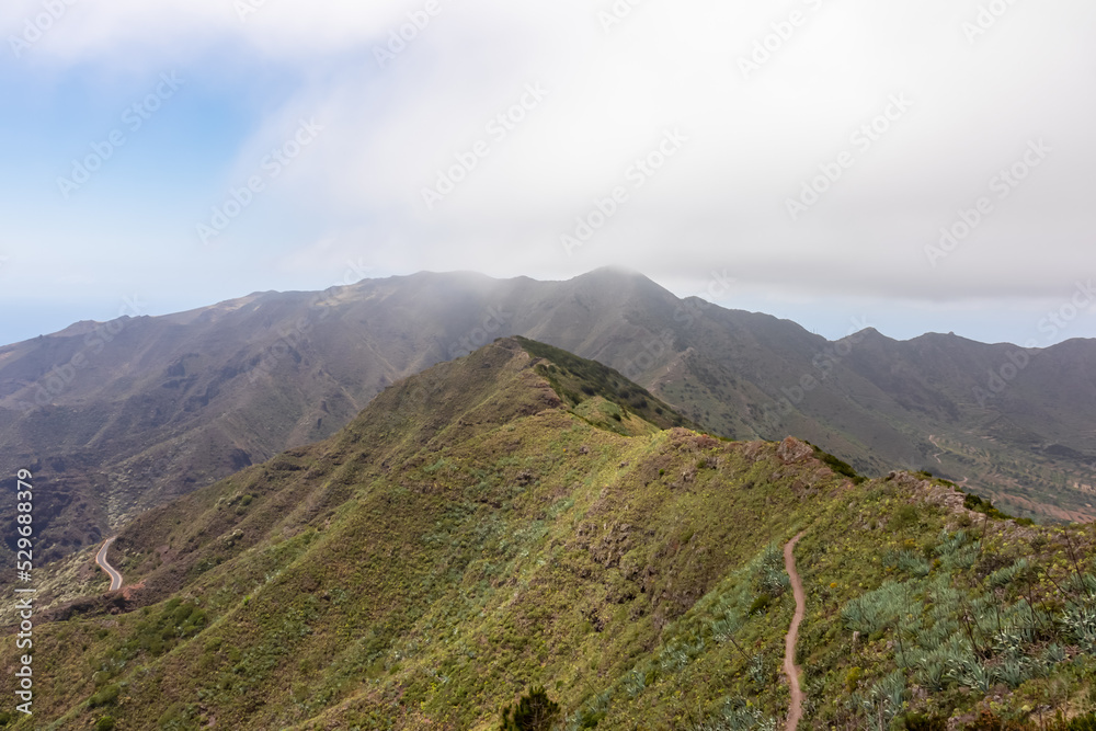 Panoramic view on scenic hiking trail over lush green hill in Teno mountain range, Tenerife, Canary Islands, Spain, Europe. Path leads to remote village Masca. Moody mystical vibes on tropical island