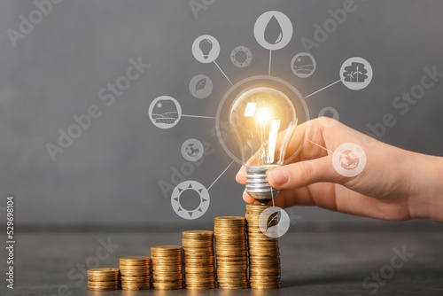 Female hand with glowing light bulb and coins on grey background. Concept of payment for electricity