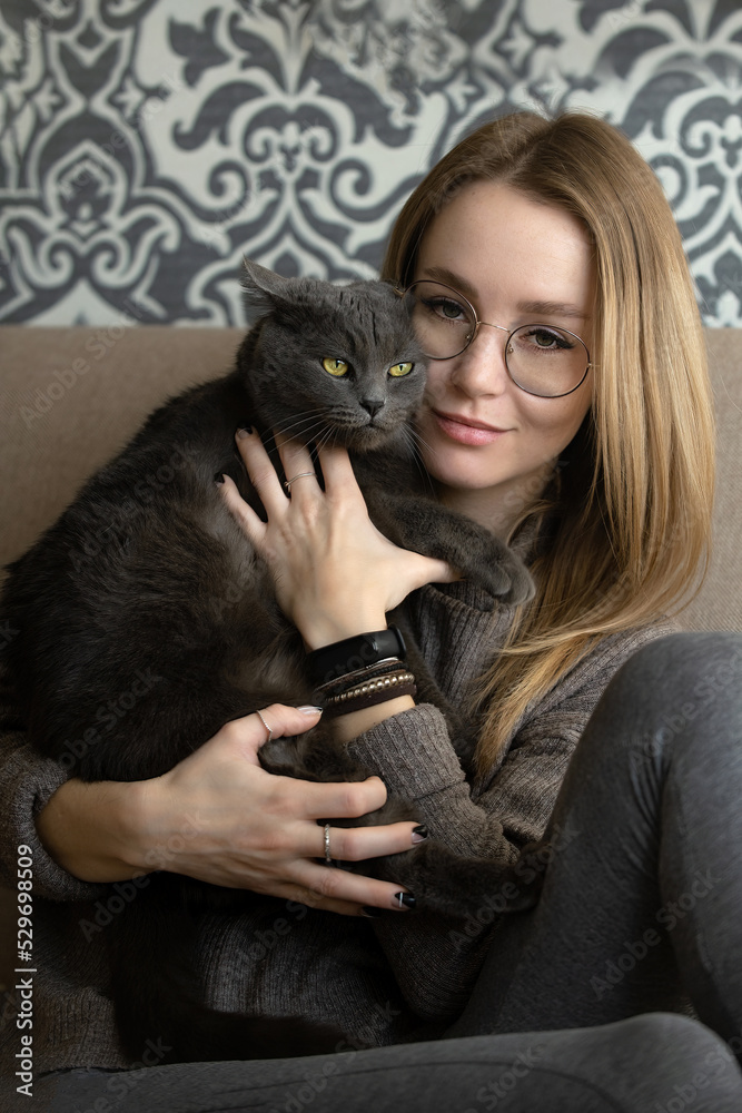 Obraz premium young beautiful woman in glasses at home on the couch holds her gray cat in her arms with a stern character and bright yellow eyes