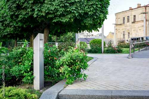 modern iron lantern of ground garden led lighting with bushes in city street with trees near flower bed landscape next to the ramp and steel railings, in the background is an old town house, nobody.