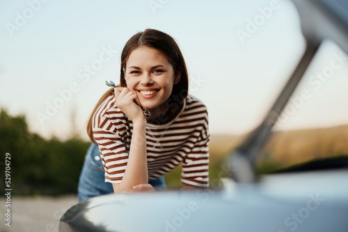 A woman with a wrench with a smile happily looks under the open hood of her car and repairs it from a roadside breakdown on a road trip alone © SHOTPRIME STUDIO