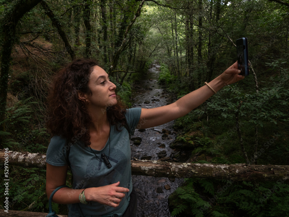 Brunette woman taking a selfie on a wood bridge with river flowing through an oak and chestnut trees in the background. Brunette woman taking a selfie on a wood bridge with river flowing through an oak and chestnut trees in the background.