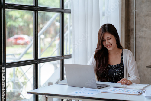 Happy young Asian businesswoman sitting on her workplace in the office. Young woman working at laptop computer in the office