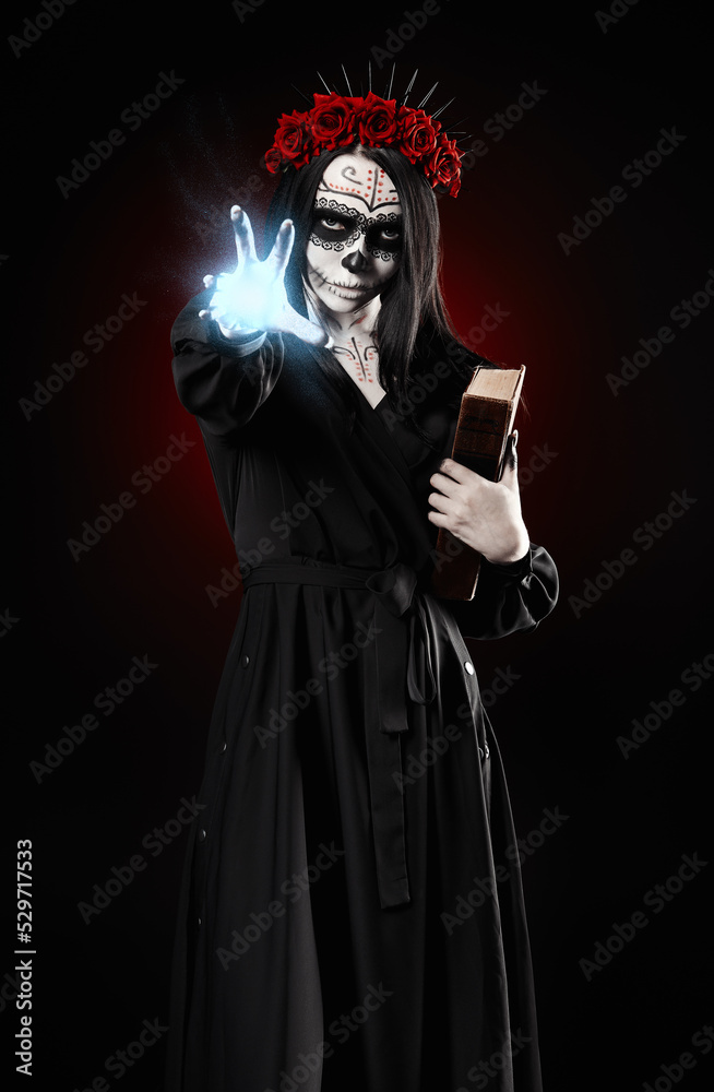 Young woman with calavera (sugar skull) makeup holds book (grimoire) and casts the spell. The witch girl with muertos (muerte) greasepaint in black dress. Day of the dead (and Halloween) theme