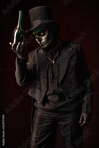 Young man in image of Baron Samedi, the Voodoo deity. Baron Saturday in black suit and top hat with a bottle of alcohol in hand. Day of the Dead (and Halloween) theme