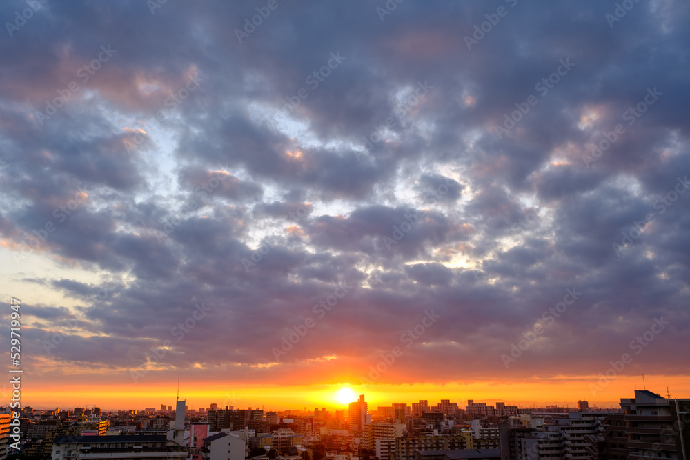都市の夜明け。日の出とともに空と雲がオレンジ色に染まり、ビル群はシルエットとして写す。神戸市街地から芦屋、大阪方面を臨む