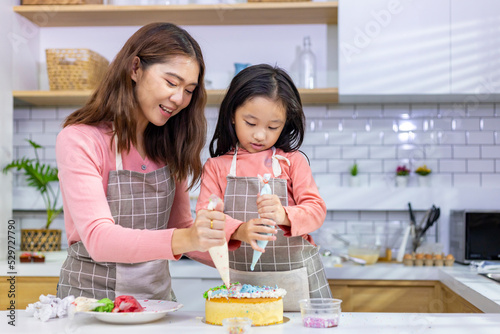 Happy Asian mother teaching her young daughter to decorating their homemade cake in white modern kitchen for home cook and baking concept