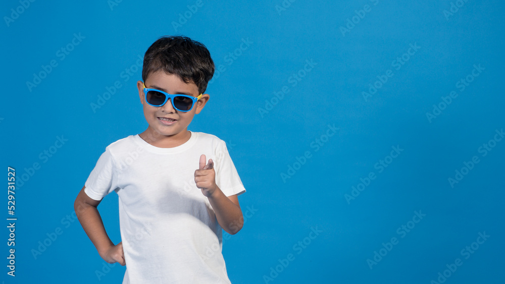 Naklejka premium Hermoso niño con playera blanca y fondo azul. Usando lentes y señalando con su dedo.