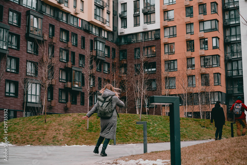 Back view of a woman in a jacket and backpack running in the modern residential building. College. Student. University. Trendy. Happiness. Walking. Walk. Vitality. Vacations. Daytime. Energy. Motion.