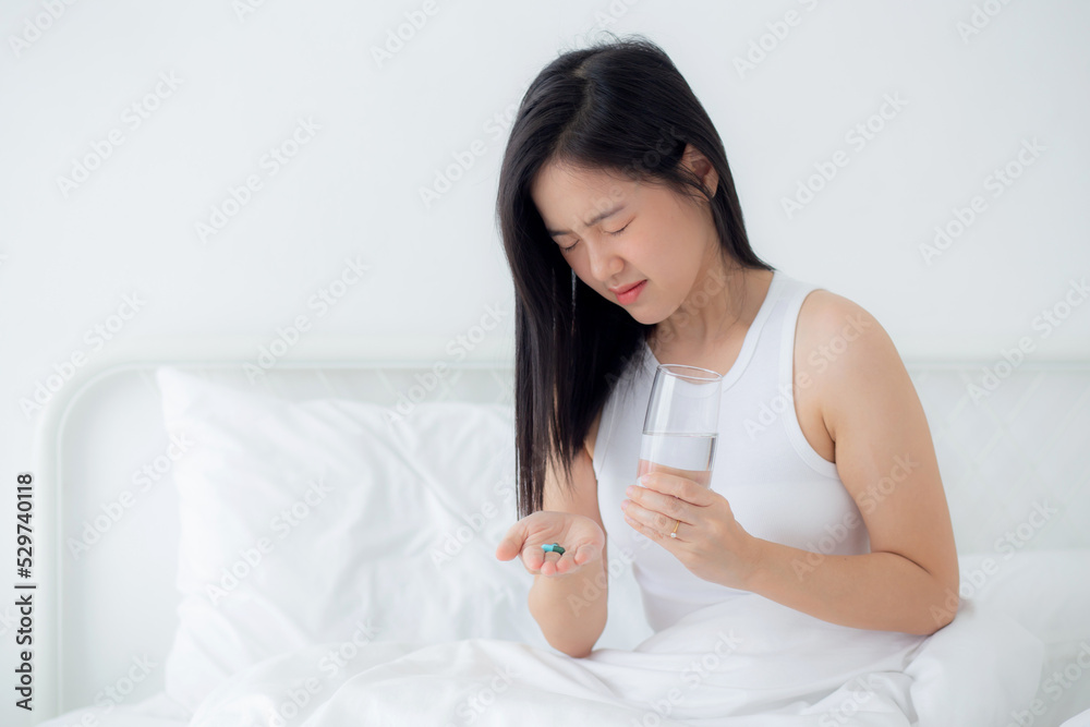 Beautiful young asian woman holding drug and glass of water while ill sitting on bed in the bedroom at home, female fever and healthcare while uncomfortable and unhappy, medical and lifestyle concept. Beautiful young asian woman holding drug and glass of water while ill sitting on bed in the bedroom at home, female fever and healthcare while uncomfortable and unhappy, medical and lifestyle concept.