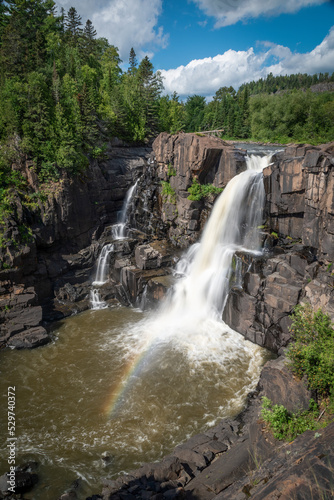 A small rainbow appears in front of High Falls in Pigeon River Provincial Park as the falls plunge down to a pool of water below. © John