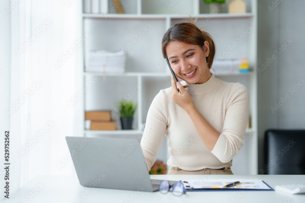 Fototapeta premium Young Asian businesswoman sit with her laptop computer happily talking on the phone with her customer explaining the detail in the office.