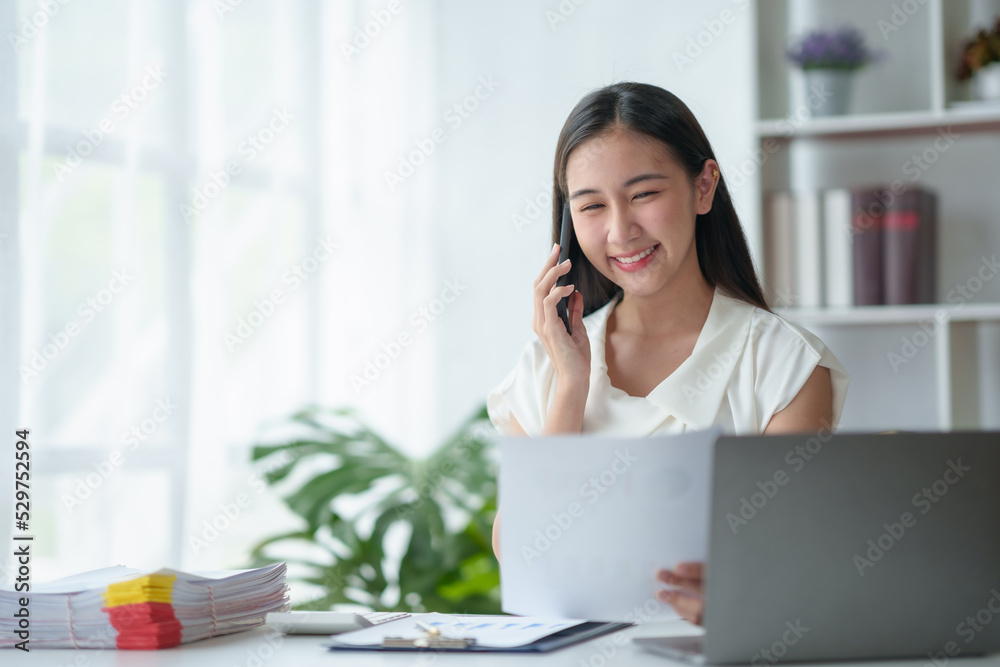 Young Asian businesswoman sit with her laptop computer happily talking on the phone with her customer explaining the detail in the office. Young Asian businesswoman sit with her laptop computer happily talking on the phone with her customer explaining the detail in the office.