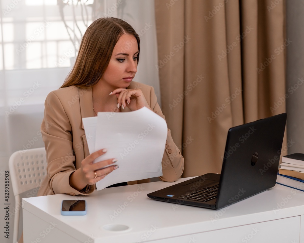European professional woman sitting with laptop at home office desk, positive woman studying while working on PC. She is wearing a beige jacket and jeans. European professional woman sitting with laptop at home office desk, positive woman studying while working on PC. She is wearing a beige jacket and jeans.