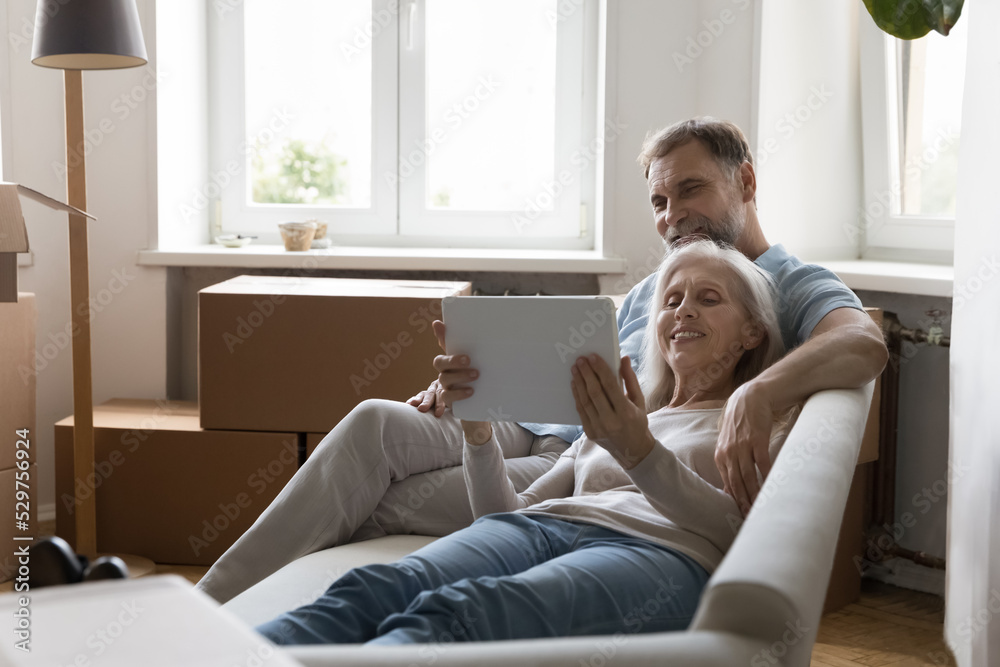 Happy retired senior family couple relaxing on sofa after moving activities, sharing tablet computer, looking at screen, laughing, making video call, using online app, Internet service