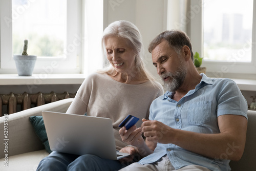 Happy middle aged elder family husband and wife shopping on Internet, paying for online purchase by credit card, using bank ecommerce app on laptop, resting on couch at home