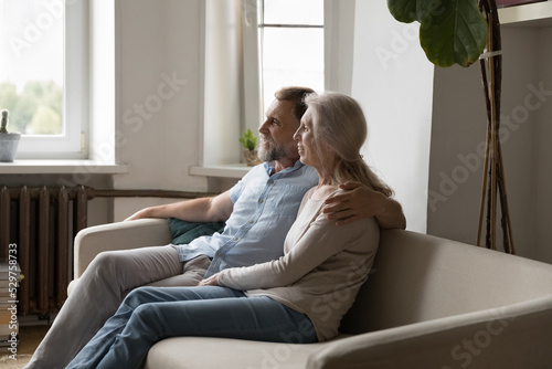 Happy mature retired family couple sitting on sofa indoors, hugging, looking away, talking, dreaming, enjoying leisure at home, hugging with love, care, affection. Retirement, relationship