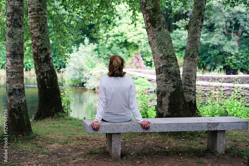 Woman rear view in a white sweater sitting alone on a stone bench in front of the river in a natural environment. Vacation and rural life concept